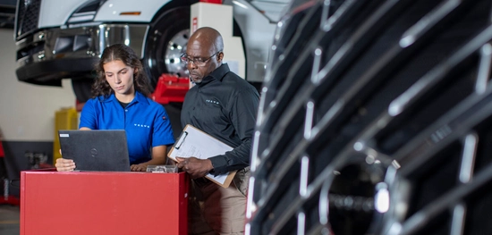 A man and a woman stand next to a tall, red desk, looking at a laptop. The man is holding a clipboard. The right side of the image is covered by a Volvo truck grill.